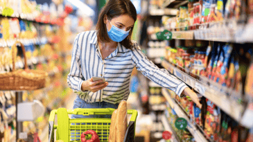 Woman with mask grabbing item off grocery shelf holding smart phone