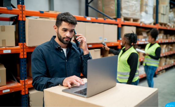 Male merchant doing stock inventory in grocery warehouse on laptop while talking on the phone