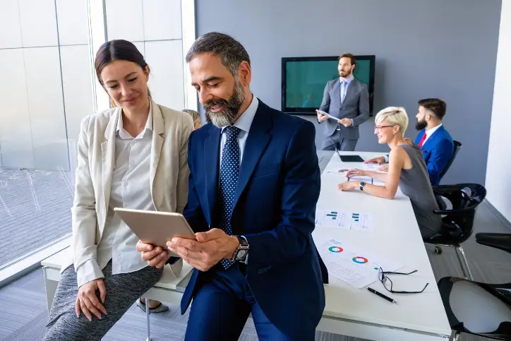 Female and male insurers looking at tablet in board room with colleagues sitting behind them