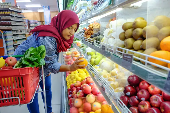 Woman wearing a hijab grabbing an orange from the produce aisle to place in her shopping basket